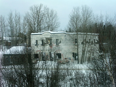 [The building is a two-story white block with a curved roofline above the middle section. The window are square and it appears paint is chipping off the building. Lots of leafless trees beside and around it as well as in the foreground.]